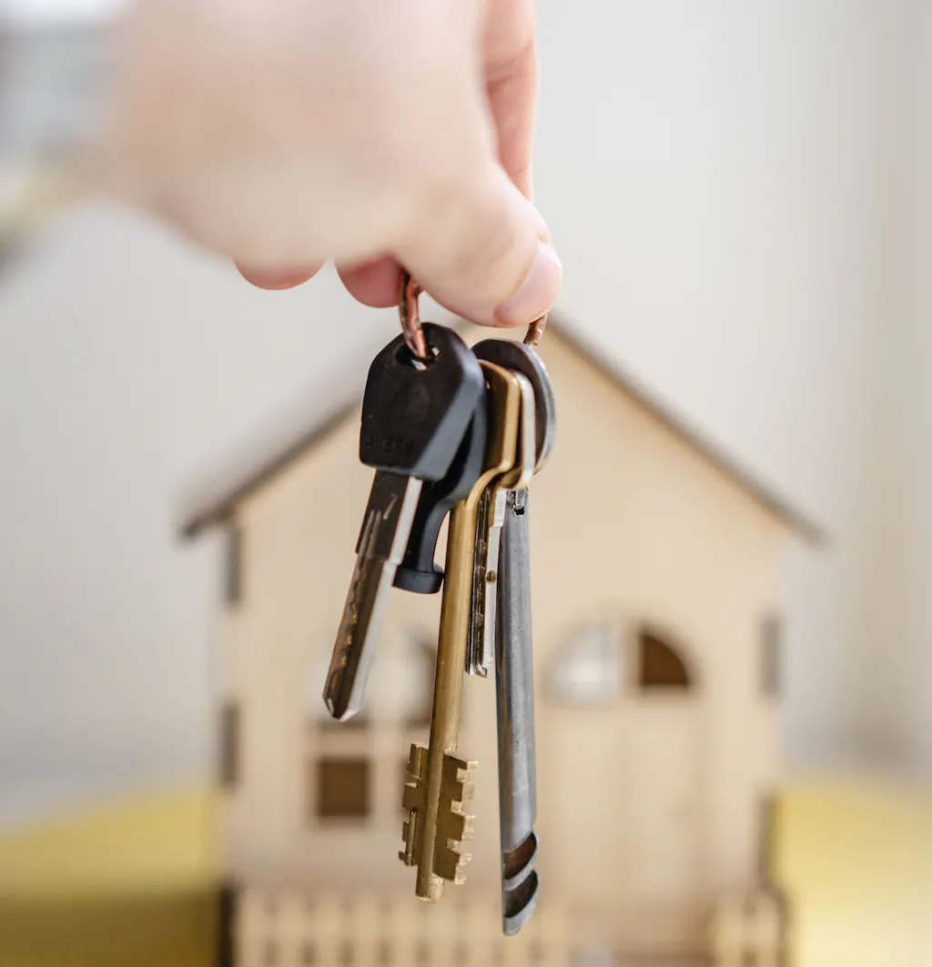 Mano sosteniendo llaves frente a una casa de madera decorativa